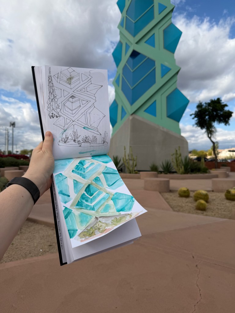 A sketchbook held open to show ink sketches and a direct watercolor painting of the Frank Lloyd Wright spire, with the actual spire visible in the background — Frank Lloyd Wright Commemorative Park, Scottsdale, Arizona.