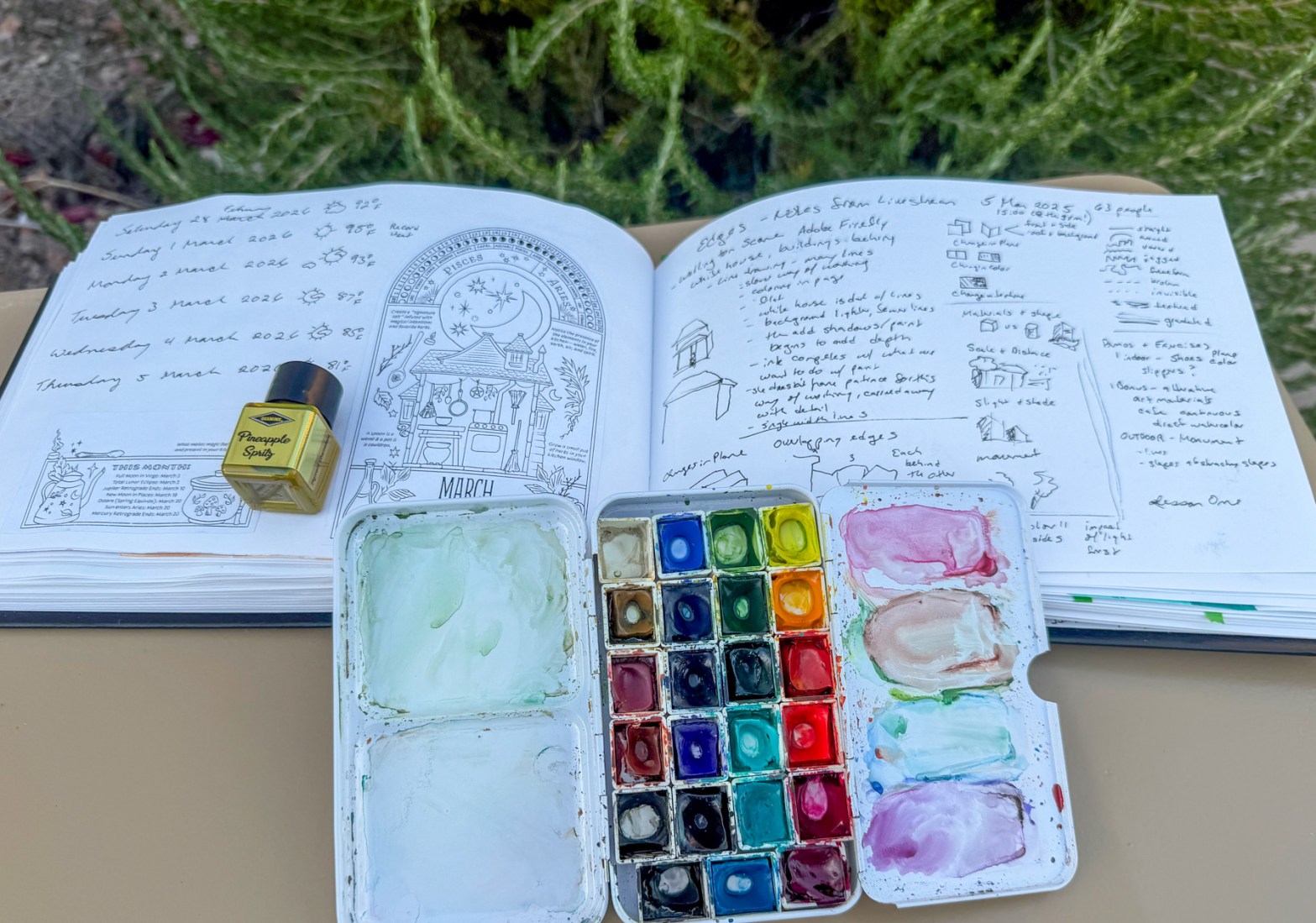 An open sketchbook showing black and white collage images, dates and weather symbols, and handwritten class notes and diagrams, with a small bottle of yellow ink and a well-used open watercolor palette resting on top.