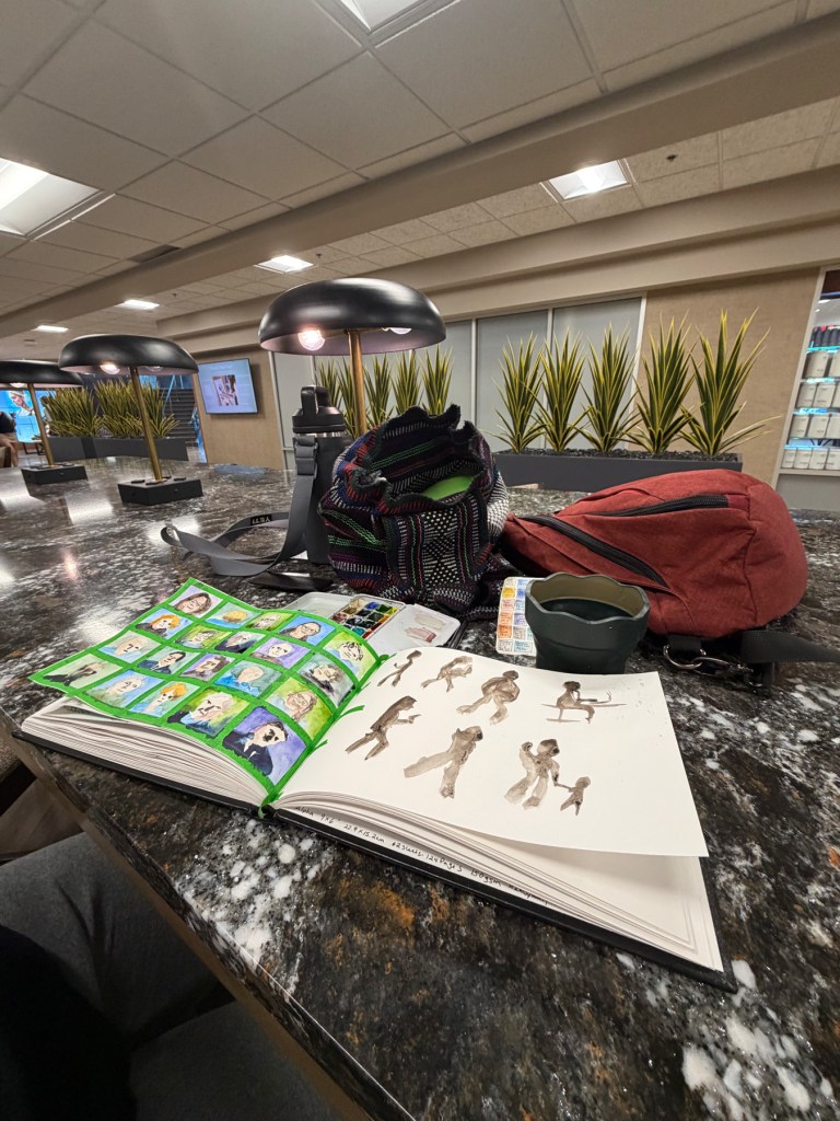 Photograph of an open sketchbook on a dark marble counter in a gym business center, showing a grid of small colorful watercolor portraits on the left page and loose sepia gesture figures on the right. A watercolor palette, woven bag, and red backpack sit nearby. The room behind has dome pendant lamps and tall spiky plants.