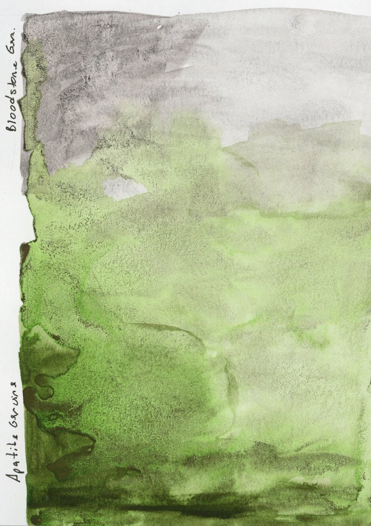A large vertical watercolor swatch on white sketchbook paper showing two pigments applied together. The upper portion is Bloodstone Genuine in a cool dark granulating grey, labeled at the top left. The lower portion transitions into Apatite Genuine, a luminous granulating grey-green, labeled at the lower left. The granulation of both genuine pigments is visible as the particles settle unevenly into the paper tooth, creating a rich textured surface. Stillman and Birn Alpha sketchbook.